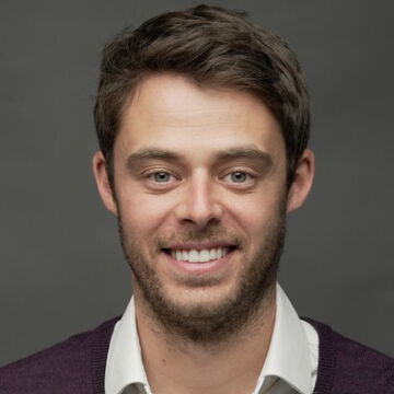 Man with dark hair and beard, smiling, wearing dark sweater over white shirt against gray background