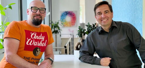 Ronny Büchner and Tino Favetto sit next to each other and smile into the camera.