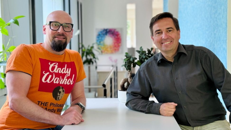 Ronny Büchner and Tino Favetto sit next to each other and smile into the camera.