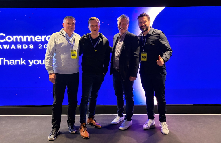 Four men wearing event badges stand smiling in front of a blue stage at a Commerce Awards event.